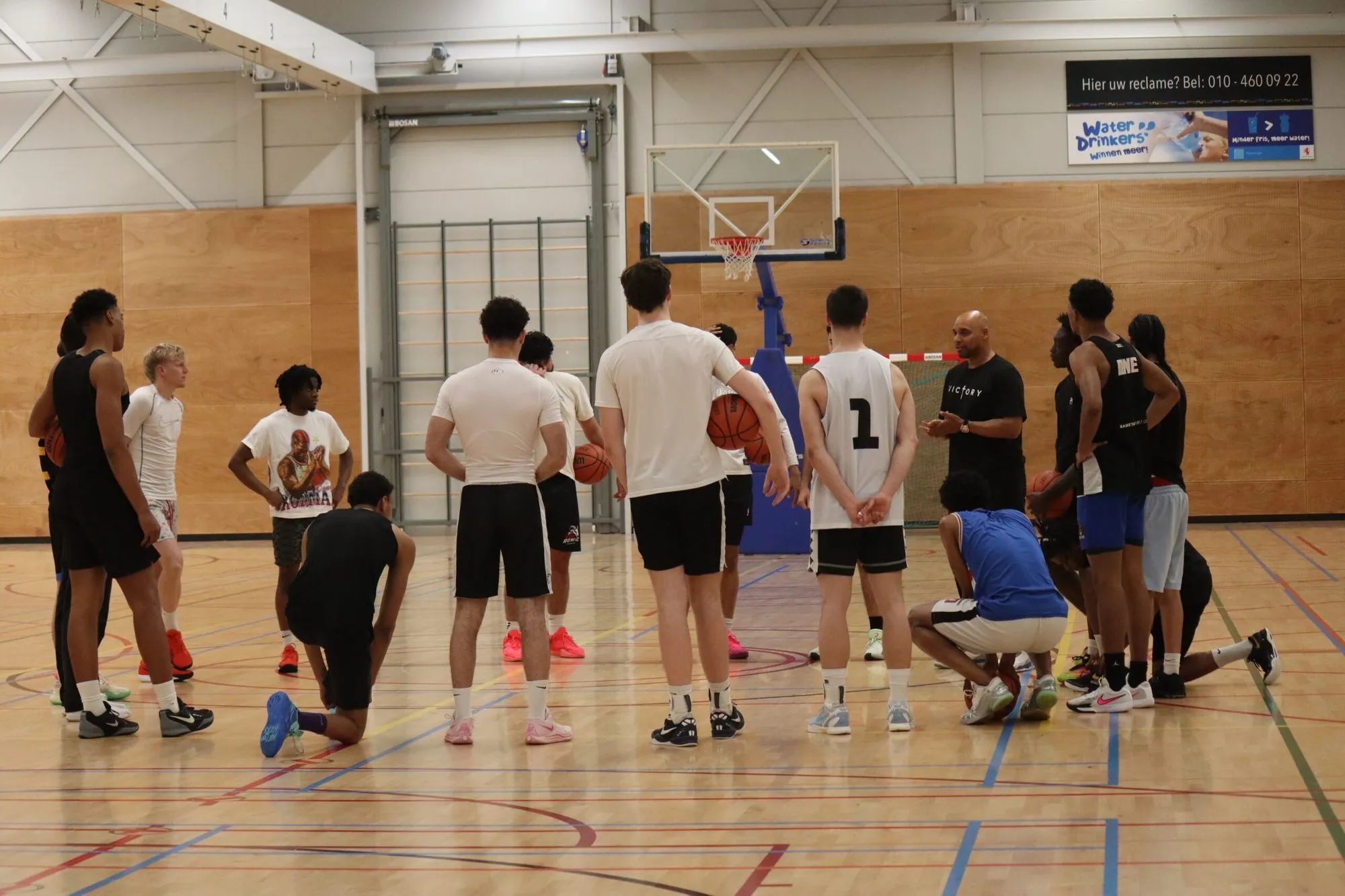 Coach van NRG Basketball geeft uitleg aan de hele ploeg, die in een kring om hem heen staat in de Erasmushal